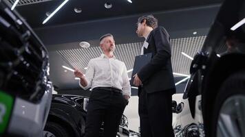 A happy middle-aged man in a white shirt communicates with a brunette guy in a business suit with a tablet in his hands by a manager in a car showroom while viewing cars video
