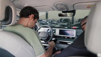 A brunette guy in a green T-shirt sits in a car with a manager in a business suit at a car dealership and communicates. Communication between a manager in a business suit and a client to guys in a car video