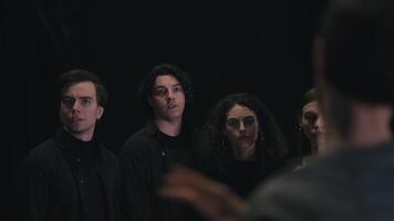 Close up, four confident young actors in black suits stand in a row and listen to their director Skip and papers and the plot on them during a rehearsal for a play in the theater video