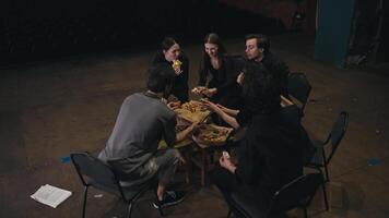 A confident small group of actors in black suits sits in a semicircle and is given pizza for lunch while preparing for a performance along with their stage director on the theater stage video