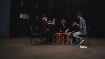 View from Zoom in of a confident group of young actors in black uniforms sitting around a pizza and taking a slice during their lunch and break while preparing for a performance in the theater video