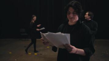 A group of actors are preparing for a performance. A young brunette man in a black suit reads out his words and scripts from sheets of paper during his preparation for a rehearsal for a performance in video