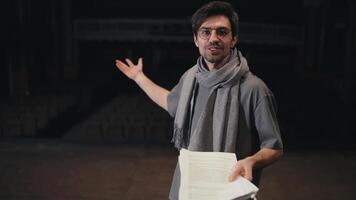 Portrait of a confident young brunette man in glasses of a confident stage director in a gray T-shirt and a gray scarf with a script in his hands on stage in the theater video