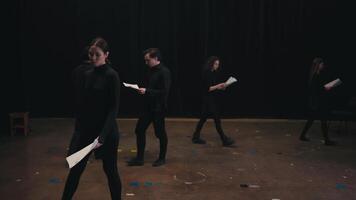 A group of confident actors in black suits walk around the stage with sheets of script and text in their hands. A brunette girl stands up straight and reads her text before the start of the video