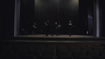 View from afar a confident group of young actors in black suits walk around the stage with sheets of paper in their minds reading their text and script of the play and preparing for their performance video