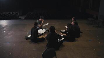 A confident young male director with script sheets in his hands tells young actors in black suits how to play their roles correctly during preparation and rehearsal for a performance on stage in a video