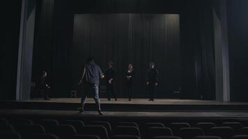 A confident stage director in a gray T-shirt comes on stage to explain to the actors how to play and move correctly on stage during preparation for a performance in an empty theater with black video