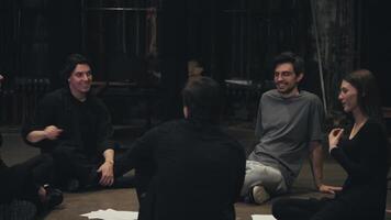 Close up a group of confident young actors in black suits together with their stage director in a gray T-shirt sit in a circle on the stage and communicate with each other during a rehearsal for a video