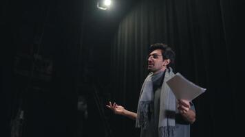 Side view of a confident male stage director in a gray T-shirt and scarf walking onto the stage along with a stack of papers and greeting everyone in the theater with black curtains video
