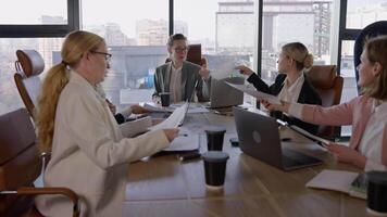 A group of businesswomen in business clothes are communicating during a meeting and passing each other sheets with graphs and ideas about their future work in a modern society with large windows video