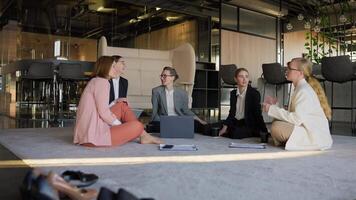 Zoom in approaching a group of confident business girls in business uniforms communicating while sitting on a soft carpet in a modern office video