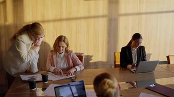 Side view of a group of businesswoman in business suits sitting at a wooden table in a sunny office communicating and working on laptops video
