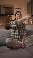 Vertical of a happy brunette girl in a plaid shirt sitting on a gray sofa and switching channels on the TV using the remote control in a modern apartment at home near her boyfriend in the video