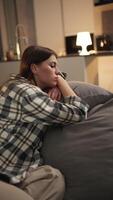 Vertical of a tired and bored brunette girl in a checkered shirt lies on the sofa leaning on the back and on the pillow in a modern apartment at home in the evening video