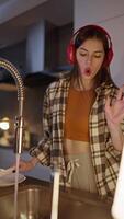 Vertical of a happy and cheerful brunette girl in a checkered shirt wearing red wireless headphones listening to cheerful and firewood music while washing dishes in the evening in a modern video