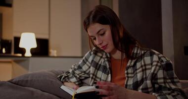 A happy brunette girl in a plaid shirt and an orange T-shirt reads a book and flips through its pages while relaxing on a gray sofa in a modern apartment in the evening video