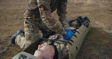 Side close view of a confident young man in camouflage uniform and blue medical gloves tying and stabilizing an unconscious man on a green military stretcher in the steppe during a training ground and video