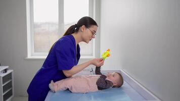 Happy brunette girl with glasses in a blue doctors uniform plays with a little girl baby during an appointment with pediatricians in a modern clinic video