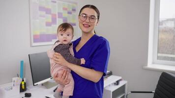 Portrait of a confident and happy brunette doctor girl in glasses and a blue uniform who is holding a little baby girl in her arms. Portrait of a confident and qualified female doctor in a blue video