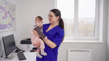A happy brunette girl in a blue uniform a doctor in glasses holds a little baby girl in her arms and finds a common language with her during prevention in a modern clinic with a pediatrician video