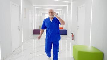 A confident doctor with a gray beard in glasses and a blue uniform hurries and walks along the corridor of a modern clinic communicating on a white smartphone video