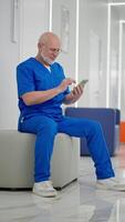 Vertical of a confident elderly man with a gray beard in blue doctors clothes sits on ottomans in the White corridor and scrolls through his feed on social networks during his break during the video