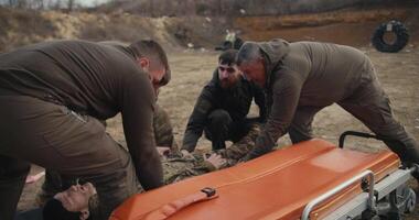 Four confident men and a soldier in a green uniform shift a training doll of a male soldier in a camouflage uniform onto an ambulance worn during training in providing medical assistance on the video