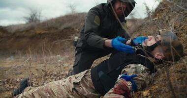 A confused male medic in a green uniform provides first aid to a wounded soldier. In a camouflage uniform and in a bulletproof vest he examines him checking his pulse and breathing during combat video