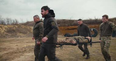 Side view of four confident male soldiers in dark green uniforms carrying a wounded doll of a male soldier in camouflage uniform on a stretcher and placing him on the ground near an ambulance on a video