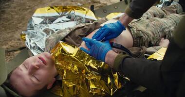 A confident male medic in blue medical gloves stops the blood of a wounded soldier in a camouflage uniform who is lying on a military medical stretcher in the combat area video