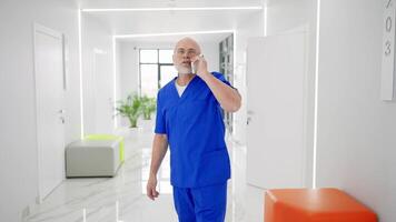 Confident male doctor of advanced age in glasses with a gray beard in a blue uniform communicates on a white smartphone and looks around while standing in the corridor of a modern clinic video