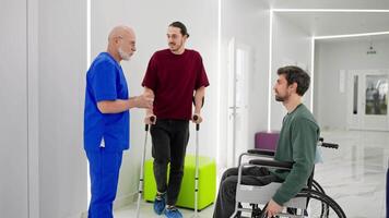 Confident man with a gray beard doctor in a blue uniform communicates with men after their injuries in a modern clinic crutches and wheelchairs for the disabled video