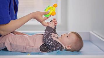 Side view of a little girl baby in a pink jumpsuit lies on a bed in a pediatrician doctors office who plays with him using a childs toy in a modern clinic for children video