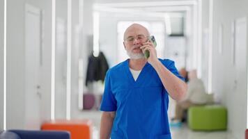 A confident elderly man with a beard a doctor walks along the corridor of a bright clinic and talks on the phone during the working day. A confident elderly doctor walks through the clinic talking video
