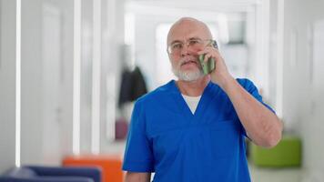 A confident man with glasses, an elderly doctor with a beard in a blue uniform walks along the corridor of a bright clinic and communicates on a green phone video