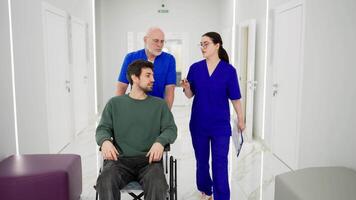 A confident elderly doctor in glasses with a gray beard and in a blue uniform takes a man to wheelchairs for the disabled while a brunette girl talks with the doctor about the rehabilitation of a video