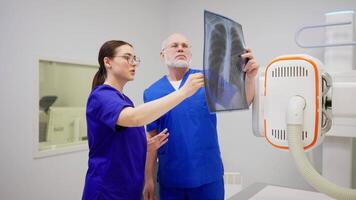 A confident brunette girl with glasses in a blue uniform communicates together and consults with a confident man doctor with gray hair while summing up the results of fluorography and confirming the video