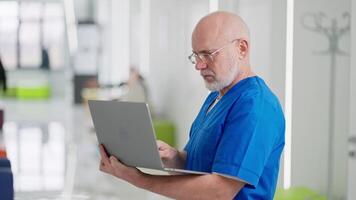 zoom in A confident elderly man with glasses a doctor in a blue uniform stands in the middle of the corridor of a bright clinic and types on his gray laptop video