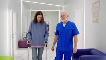 A confident doctor with a gray beard glasses and a blue uniform walks along the corridor of a modern clinic and talks to a brunette girl in a blue jacket who walks on crutches after her injury video