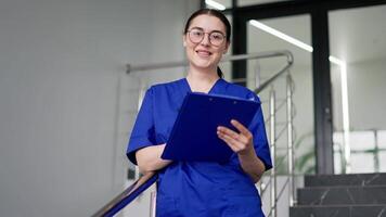 Portrait of a confident and happy brunette doctor girl in round glasses in an autumn uniform who is holding a blue tablet in her hands smiling and posing in a modern clinic on the stairs video