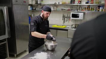 A confident male professional chef in a black uniform transfers dough from a bowl to the table and begins to knead it before preparing pizza in a restaurant kitchen video