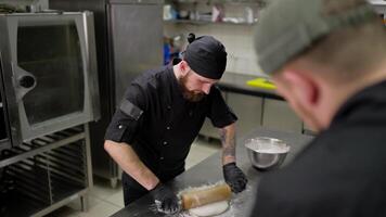 Professional male chef in black uniform with bandana rolling out dough in kitchen using dough and bowls. Confident male chef rolls out dough using a rolling pin and tells a trainee how to roll it out video