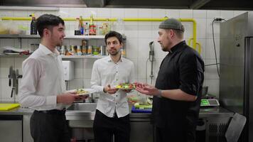 Side view of a professional chef in a black uniform communicating with two waiters in white shirts holding ready-made dishes about solving problems in the kitchen and improving dishes and their video