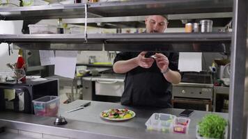 A confident male cook in a black uniform stands in the kitchen while issuing orders lays out stickers with written dishes and gives out the required dish to the hall for the waiters to hand over to video