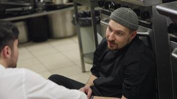 Communication between a professional cook in a black uniform and a waiter during a short break in the kitchen while sitting on the floor leaning on the table during a busy working day video