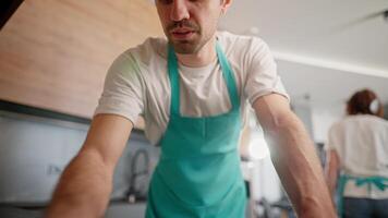An inside view from the camera a brunette guy with stubble in a white T-shirt and a blue apron washes the stove in a modern kitchen while using a cleaning company video