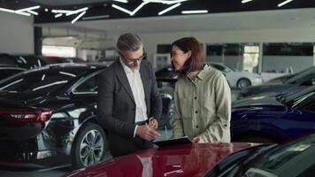 A confident middle-aged man in glasses and a business suit a manager at a car dealership communicates with a brunette girl while signing a contract regarding a car in a modern car dealership video