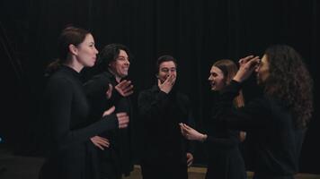 A group of happy and confident theater university students in black suits stand on stage near a black curtain and expressively communicate with each other video