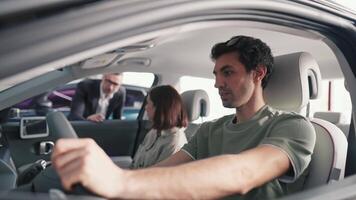A confident brunette guy in a green T-shirt sits in the car with his girlfriend and listens to a consultation from the manager about this brand of car in a car showroom video