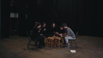 Happy actors in black suits sit around a small makeshift table with pizza and eat it during their break during a rehearsal on stage for a play in the theater video